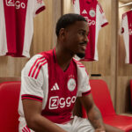 Smiling man in a red and white Ajax jersey sits on a red bench in a locker room, with hanging jerseys behind him.