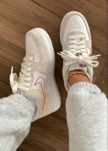 Pair of white sneakers with an orange swoosh on a wooden floor, worn with light gray sweatpants.