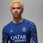 Young man with bleached blond curls wearing a blue Paris Saint-Germain jersey, facing camera in a neutral studio backdrop, sponsor logo visible.