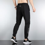 Back view of a person wearing black athletic joggers with a reflective stripe on the right leg and black sneakers, showing a slim-fit design and branded waistband composition.