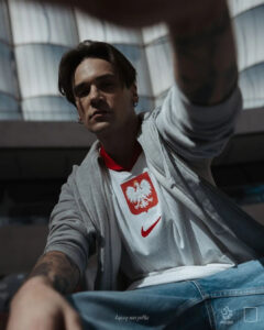 Young man wearing a white Poland football jersey with eagle crest, gray hoodie, taking a low-angle selfie outdoors.