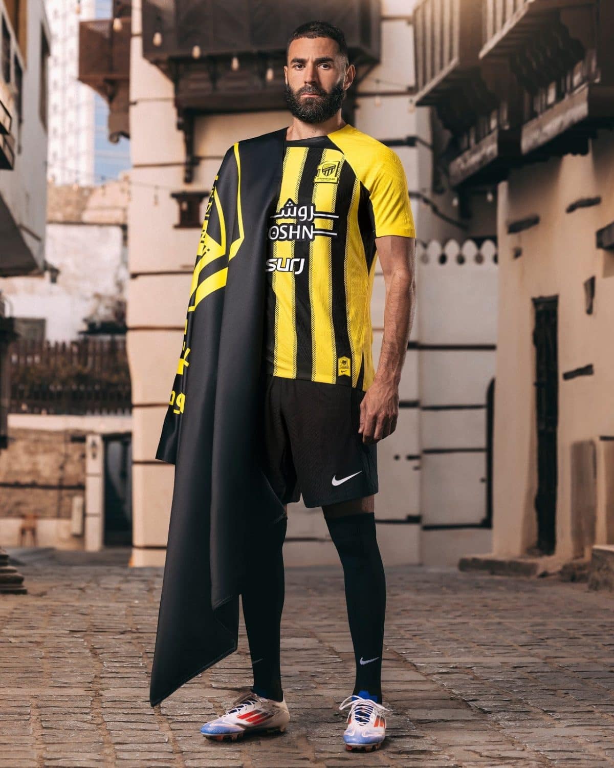 Man wearing a yellow-and-black striped sports jersey with a black cape draped over one shoulder, standing in a narrow street.