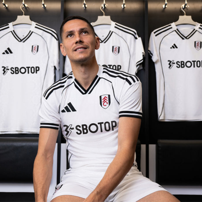 6y | UAE SPORT STORE Man sitting in a locker room wearing a white Fulham football jersey with black trim and SBOTOP sponsor, with jerseys behind him.