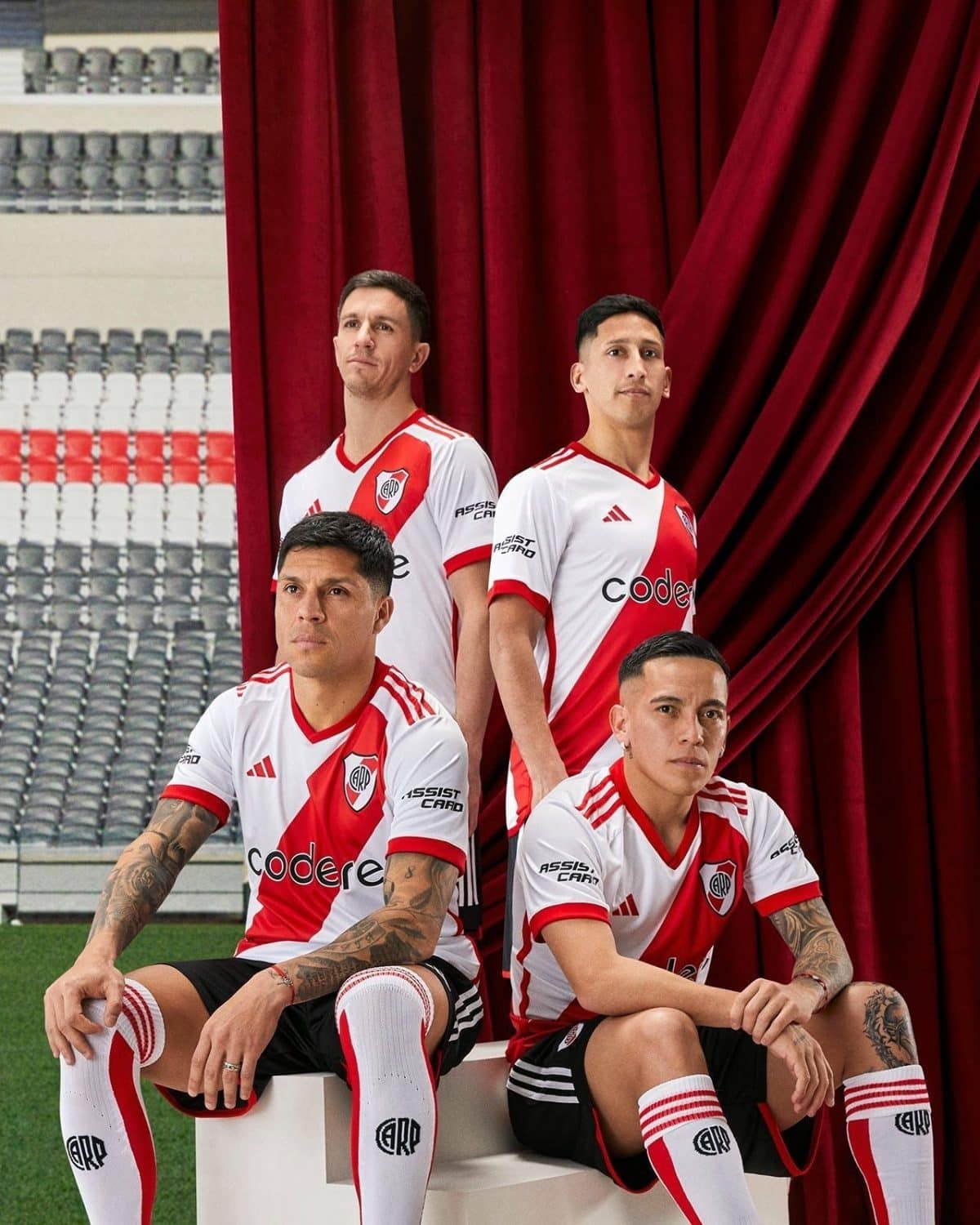Four soccer players in white and red jerseys stand and sit beside a red stage curtain, stadium seating visible in the background.