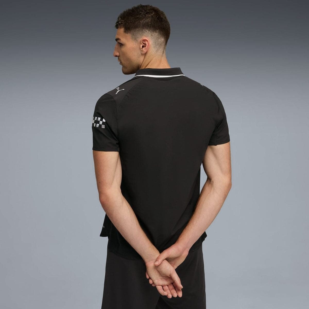 Back view of a man wearing a black polo with white collar trim and a checkerboard patch on the left sleeve, against a gray gradient background.