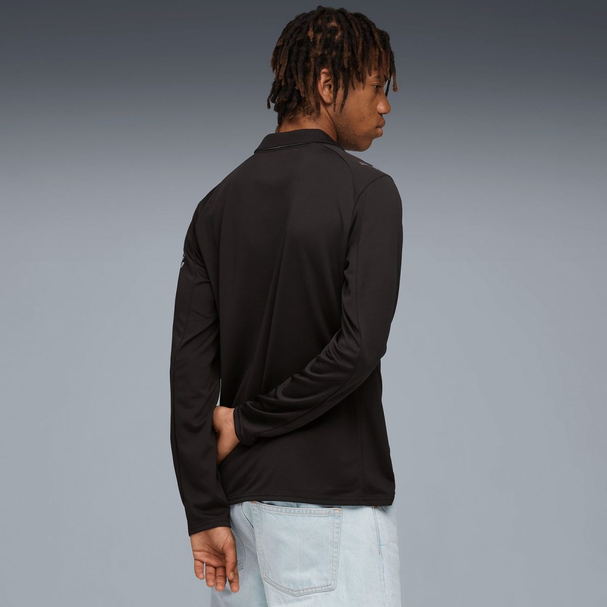 Person standing with their back to the camera, wearing a black long-sleeve shirt and light jeans, arms loosely crossed behind the back against a gray gradient background.