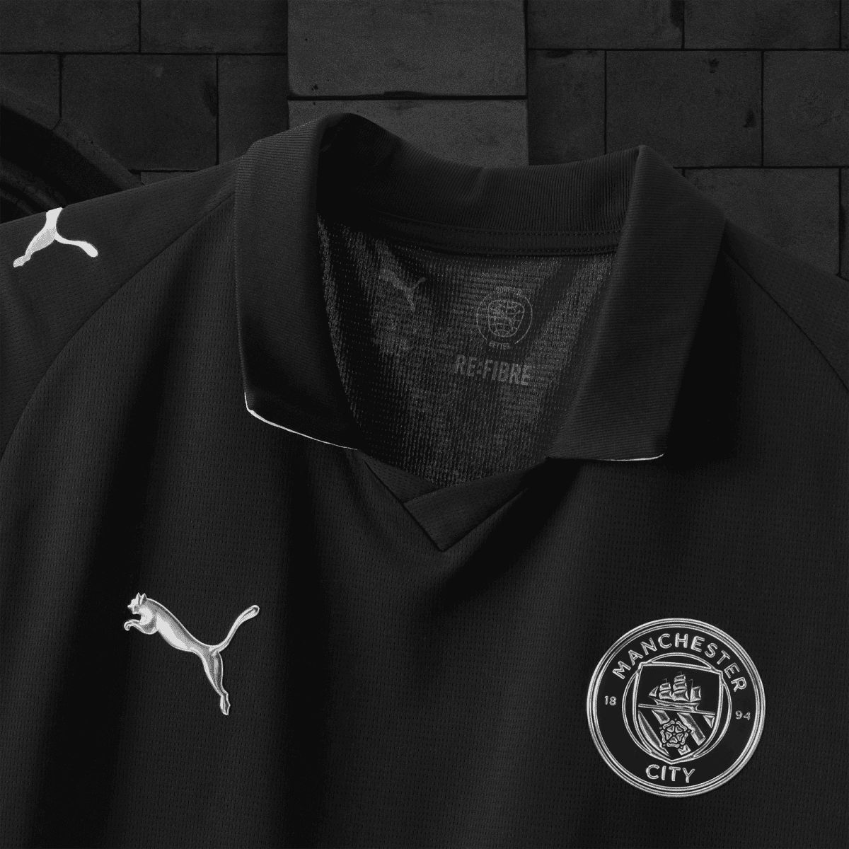 Close-up of a black Manchester City football jersey showing the white Puma logo on the left chest and the club badge on the lower right, with the collar and textured fabric visible.