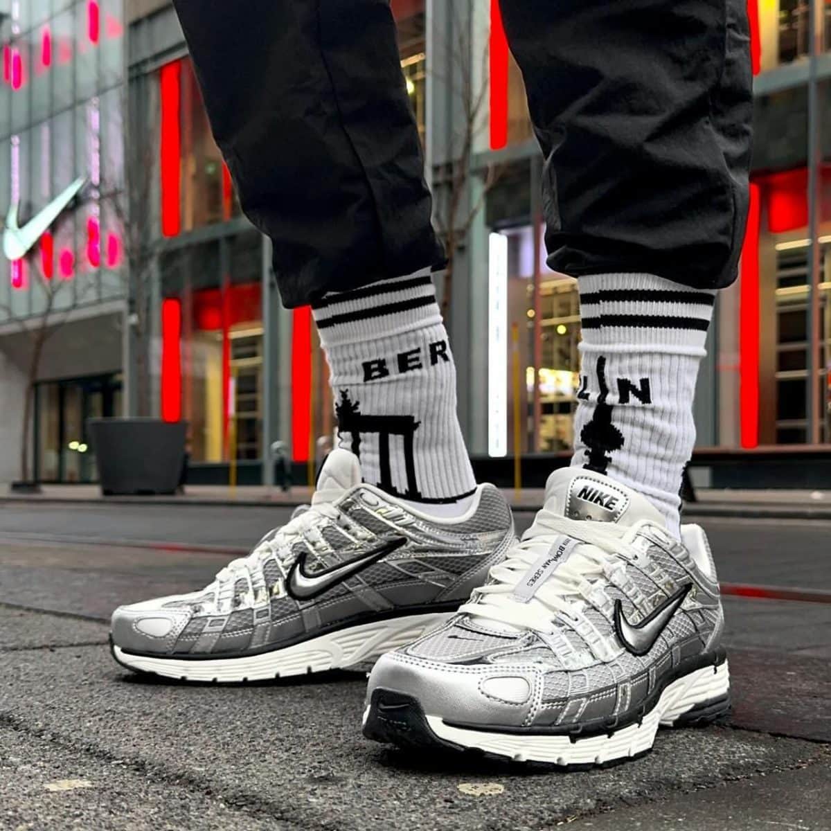 Close-up of silver and white Nike sneakers on a wet city street, paired with white crew socks featuring black stripes and the word BERLIN, with red neon lights in the background.