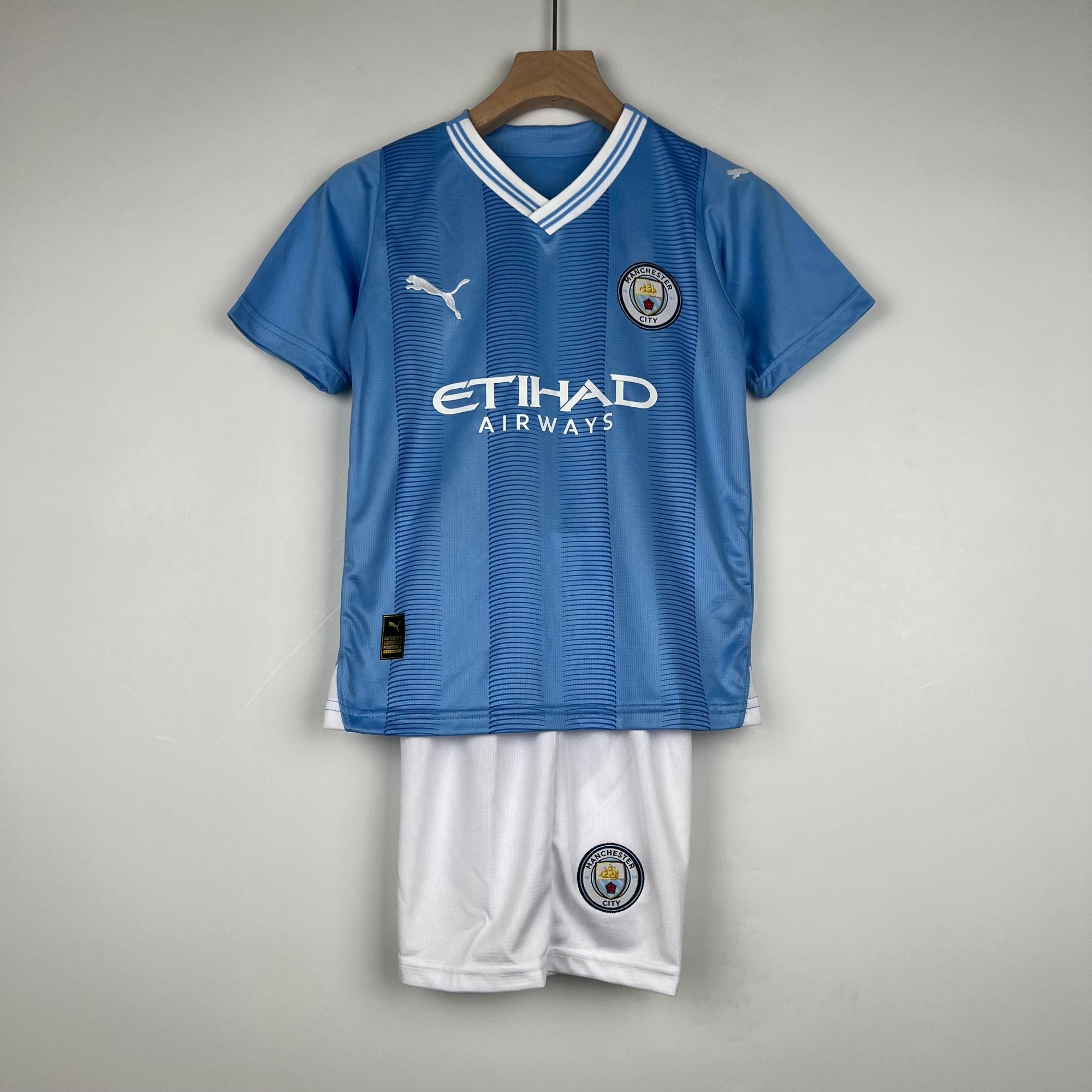703b39ac_a9363424-dc37-4ccc-9829-1768a70d97d8 | UAE SPORT STORE Blue Manchester City football jersey on a wooden hanger, with crest and Puma logo visible, paired with white shorts below