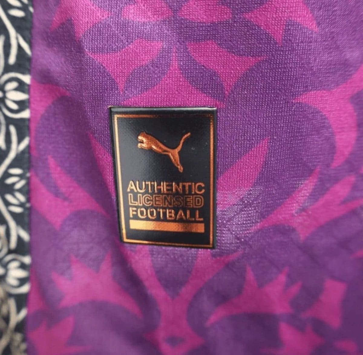 Small black tag with a gold Puma logo and the words 'AUTHENTIC LICENSED FOOTBALL' sewn onto purple patterned fabric.