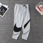 Gray Nike joggers with a large black swoosh on the left leg, drawstring waist, and cuffed ankles on a dark wooden floor.