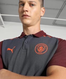 Young man wearing a dark gray Manchester City polo with maroon sleeves, seen from a low angle indoors.