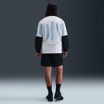 Back view of a person wearing a white jacket with light blue vertical stripes, black sleeves, black shorts, and black sneakers in a studio setting.