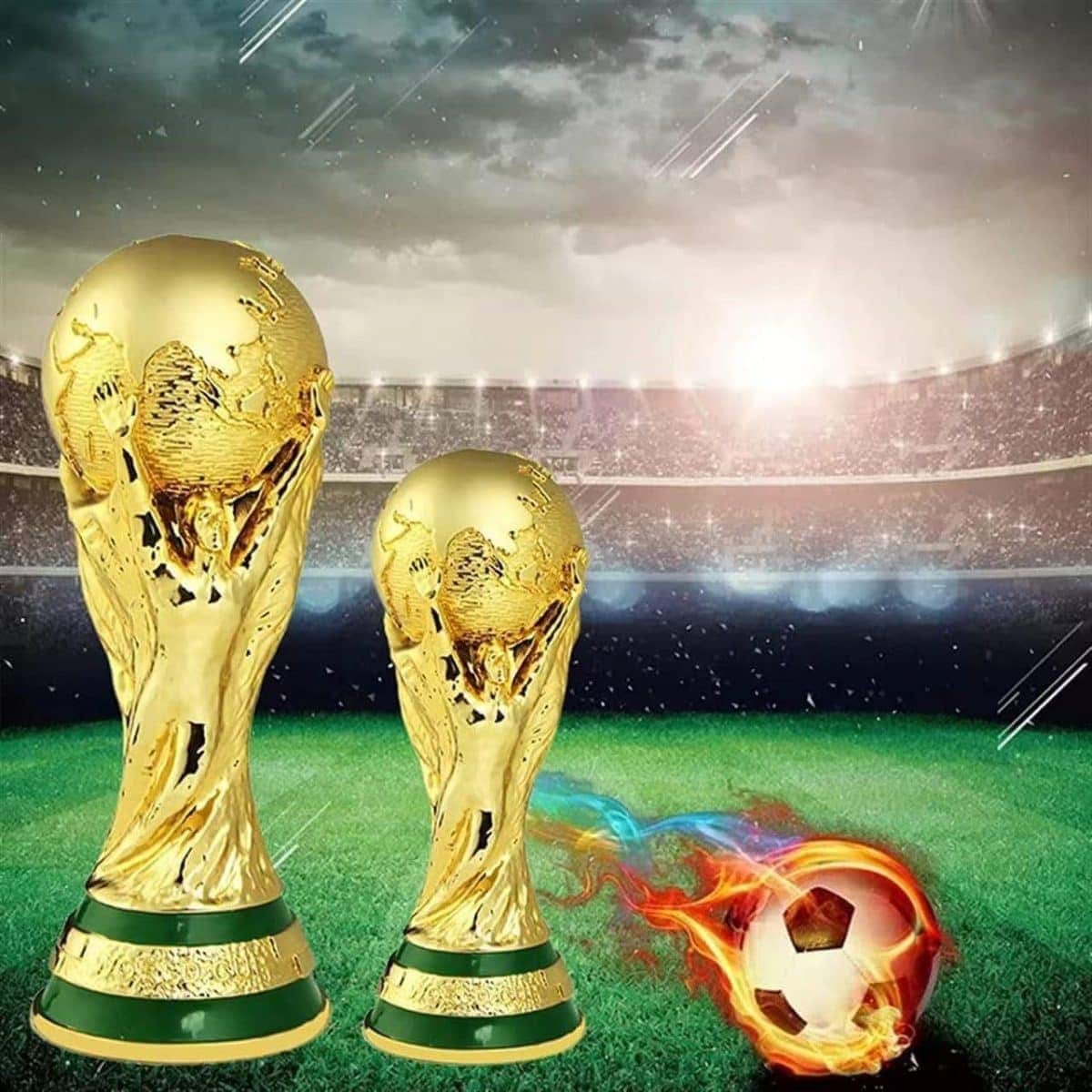 Two large gold World Cup-style trophies on a grassy soccer field with a glowing stadium in the background and a flaming soccer ball nearby