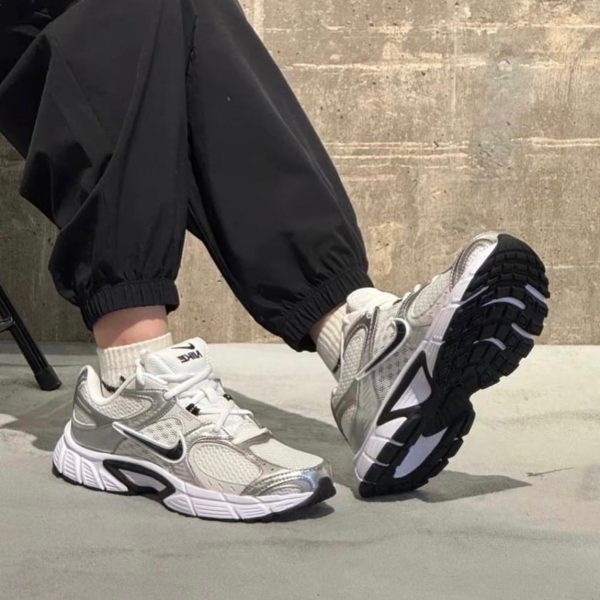White and gray Nike sneakers with a black swoosh on a person wearing black joggers, raised foot against a concrete backdrop.