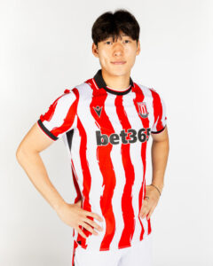 Young man wearing a red-and-white striped Stoke City football kit with the bet365 sponsor, standing with hands on hips.
