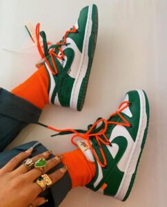 Pair of green and white sneakers with bright orange laces, worn with orange socks and multiple rings on the hand holding them.