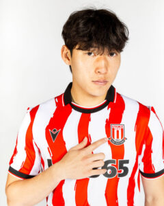 Young man wearing a red-and-white striped Stoke City football jersey points to the club badge on his chest.