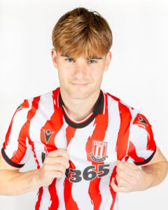 Young man wearing a Stoke City red-and-white striped football jersey, smiling at the camera with the shirt partially pulled to show the sponsor logo.