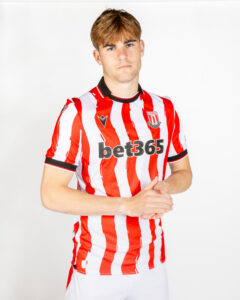Young man in a red-and-white striped Stoke City football kit posing with hands clasped in front of him, neutral expression.