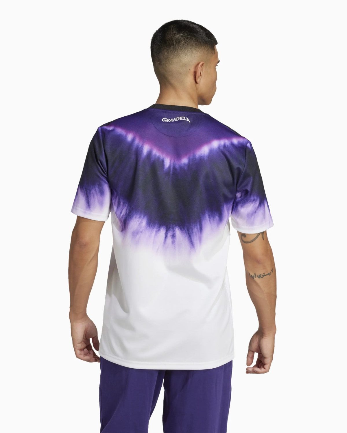 Back view of a man wearing a purple-to-white tie-dye T-shirt with a 'GRANDEZA' logo at the neckline and a visible forearm tattoo on the right arm.
