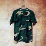 Green camouflage short-sleeve shirt on a hanger against a textured wall.