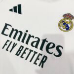 72d50d04-092d-4216-82d2-cef66e6a5b07 | UAE SPORT STORE Close-up of a white Real Madrid football jersey showing the Emirates Fly Better sponsor text and club crest on the right shoulder area.