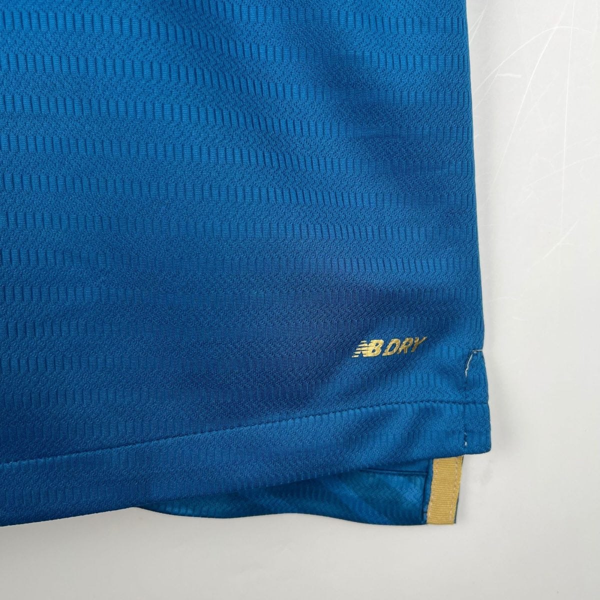 Close-up of a blue textured bag fabric with a yellow strap visible at the bottom-right and a partial New Balance logo.