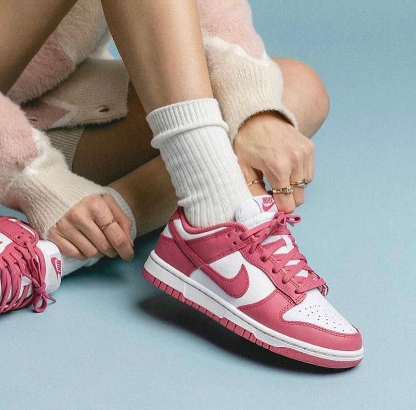 734cfc76-391f-4a08-b33d-cd919d2f95fc | UAE SPORT STORE Close-up of a person tying pink and white Nike sneakers with white socks against a light blue background.