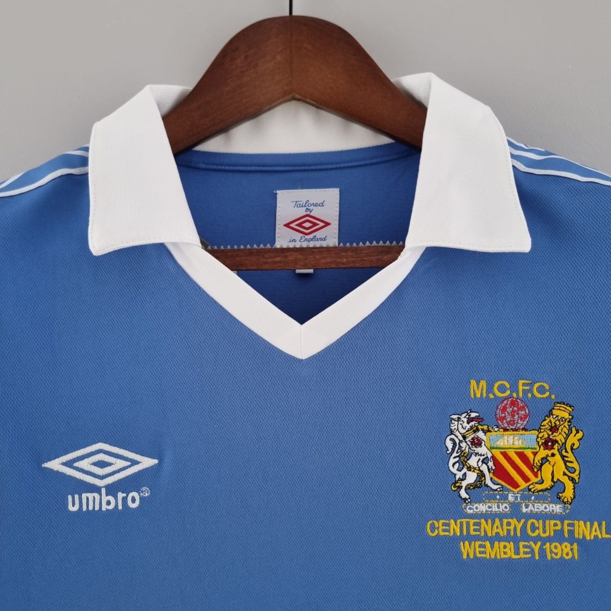Blue soccer jersey on a wooden hanger with white collar; features the Umbro logo on the left and a club crest with 'MCFC' and 'Centenary Cup Final Wembley 1981' text on the right.