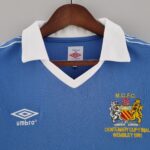 7376cc73_a53c7e32-c85f-42a0-b73b-7967e643a259 | UAE SPORT STORE Blue soccer jersey on a wooden hanger with white collar; features the Umbro logo on the left and a club crest with 'MCFC' and 'Centenary Cup Final Wembley 1981' text on the right.