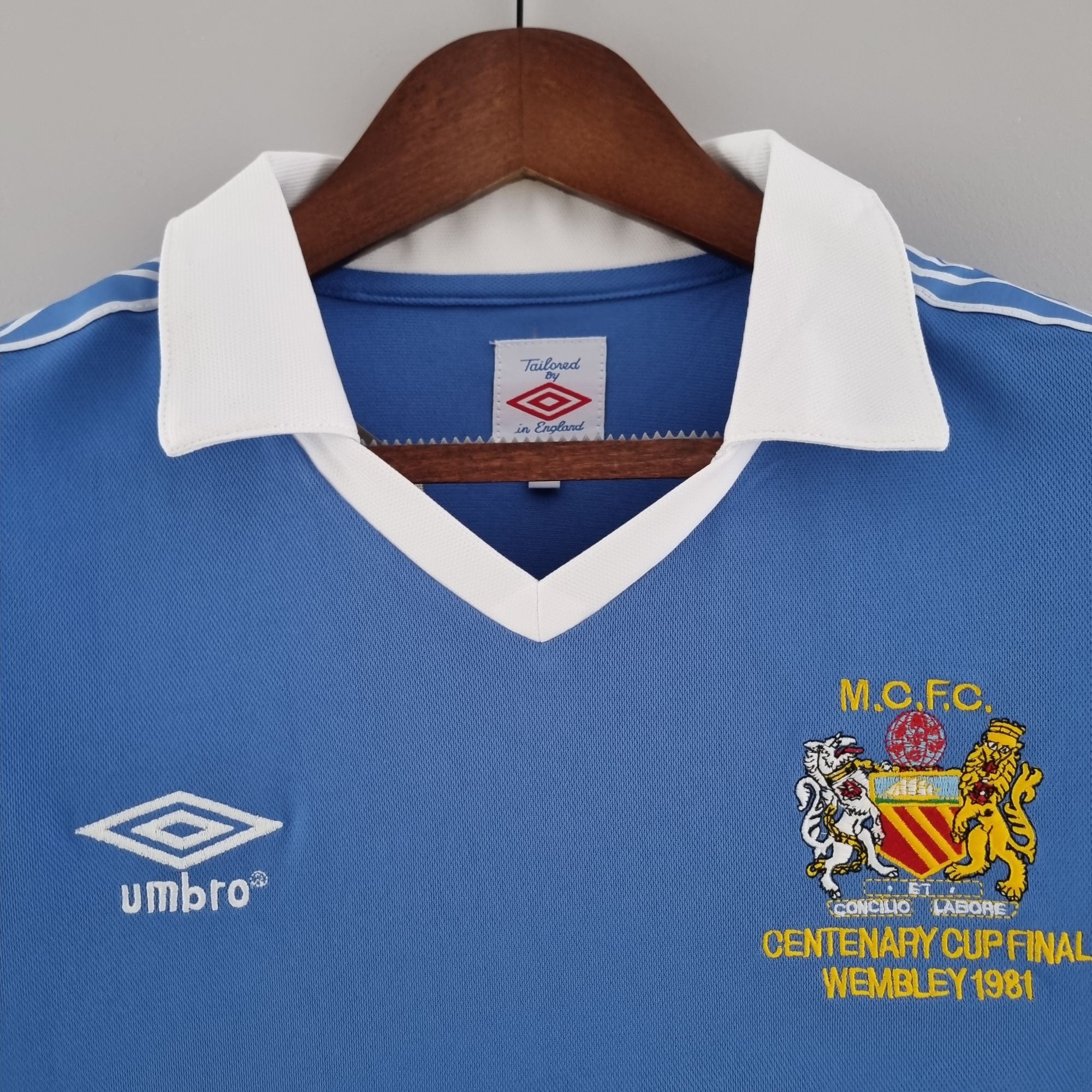 7376cc73_a53c7e32-c85f-42a0-b73b-7967e643a259 | UAE SPORT STORE Blue soccer jersey on a wooden hanger with white collar; features the Umbro logo on the left and a club crest with 'MCFC' and 'Centenary Cup Final Wembley 1981' text on the right.