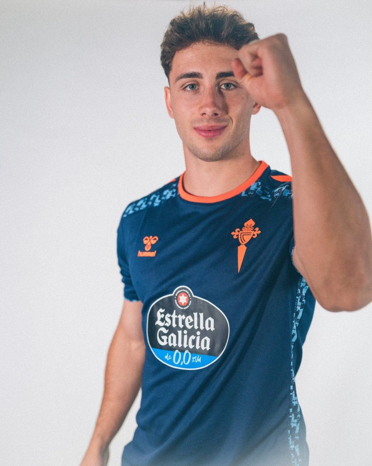 Young man in a blue soccer jersey raises his right fist in front of him, looking at the camera.