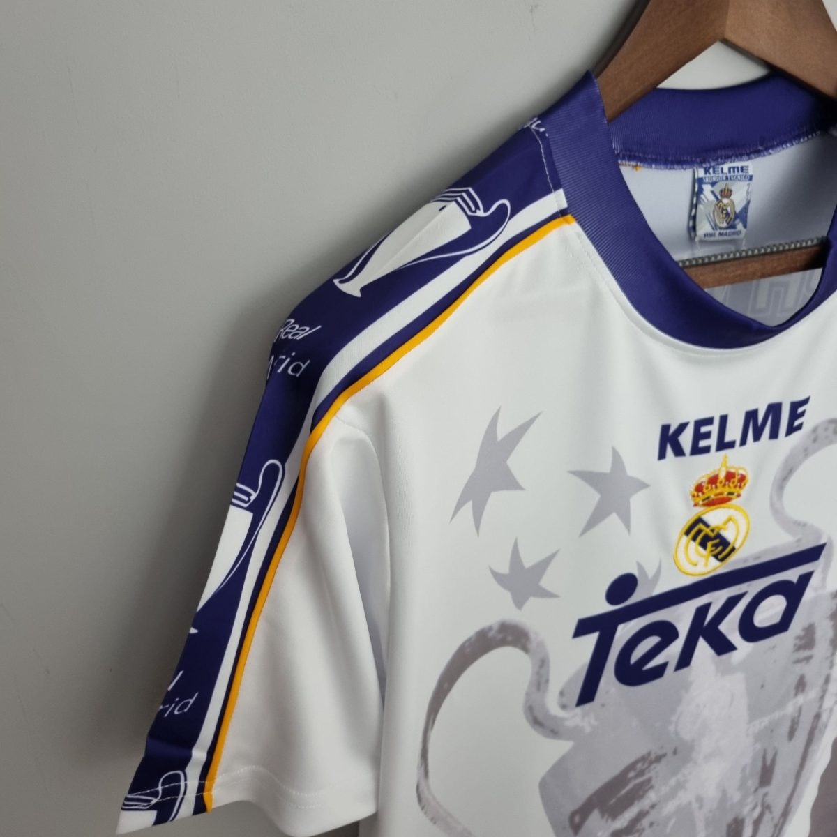 Close-up of a white Real Madrid jersey on a wooden hanger, showing the Real Madrid crest and Kelme branding.