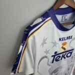 73c7c1e7_fd0985b2-38e6-4f40-8320-4853b05571a1 | UAE SPORT STORE Close-up of a white Real Madrid jersey on a wooden hanger, showing the Real Madrid crest and Kelme branding.