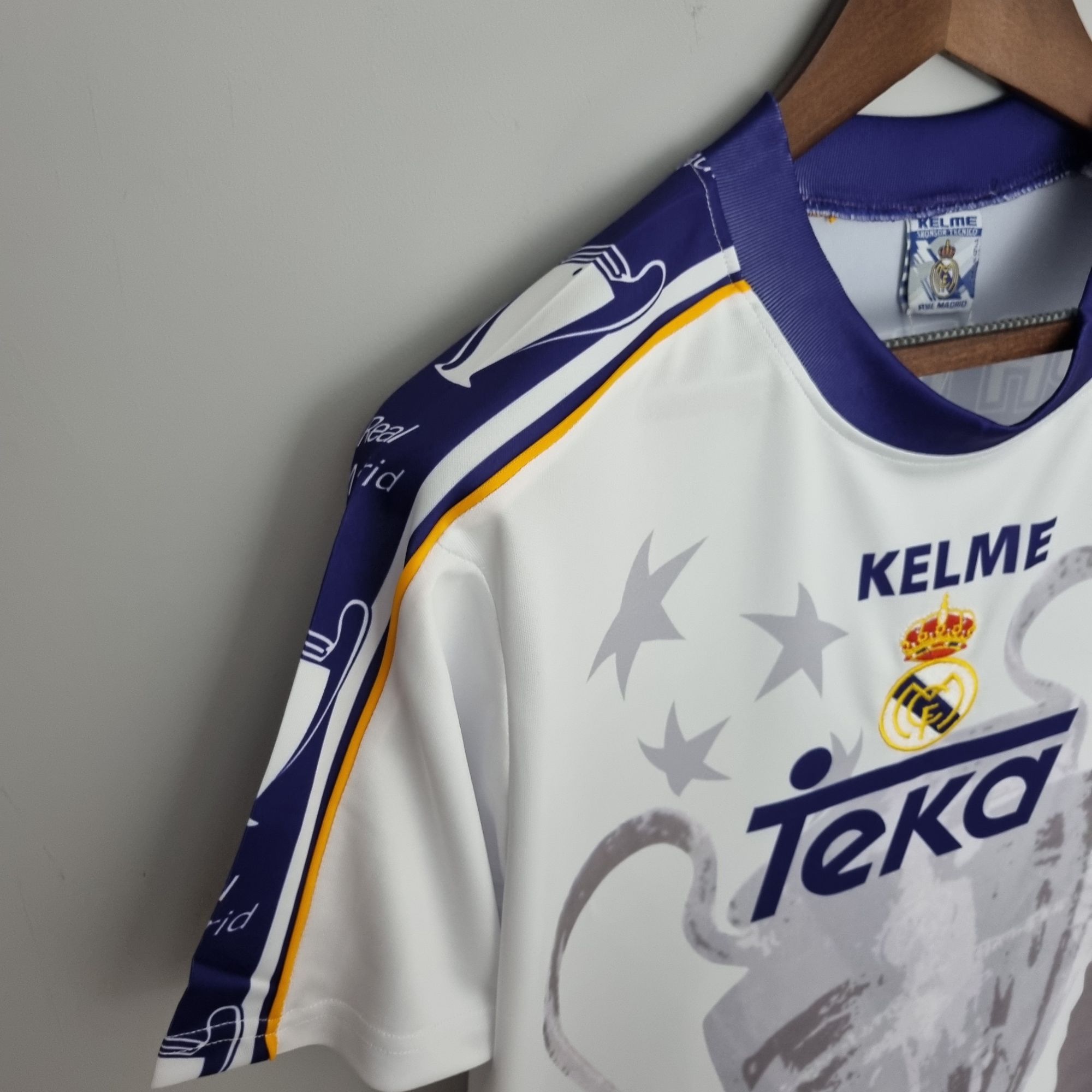 73c7c1e7_fd0985b2-38e6-4f40-8320-4853b05571a1 | UAE SPORT STORE Close-up of a white Real Madrid jersey on a wooden hanger, showing the Real Madrid crest and Kelme branding.