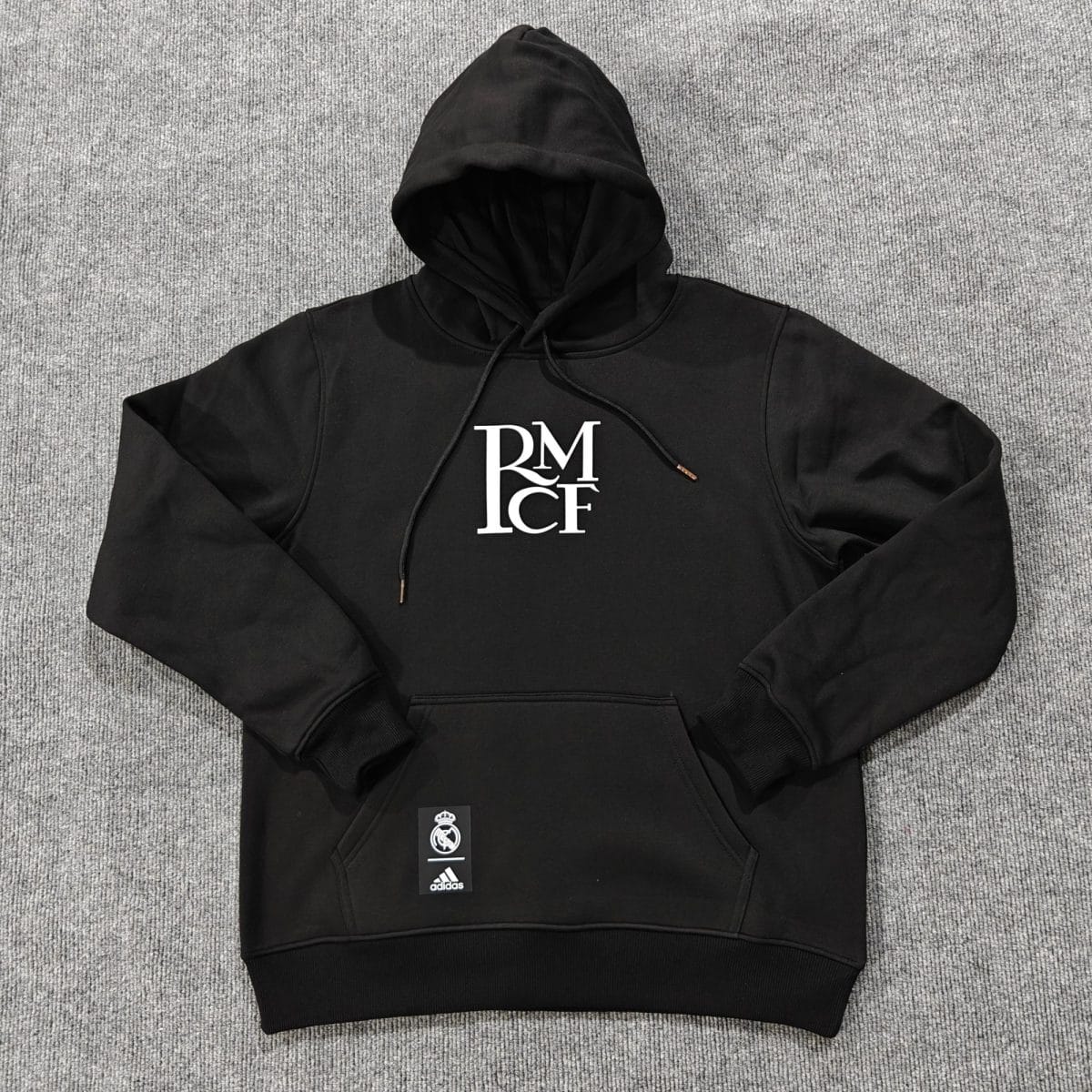 Black hoodie with hood up, white RM graphic on chest, laid on gray carpet; Real Madrid and Adidas tags attached.