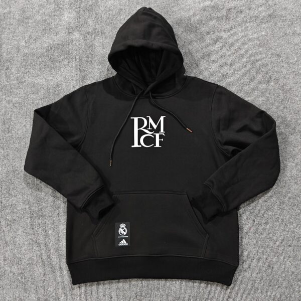 Black hoodie with hood up, white RM graphic on chest, laid on gray carpet; Real Madrid and Adidas tags attached.