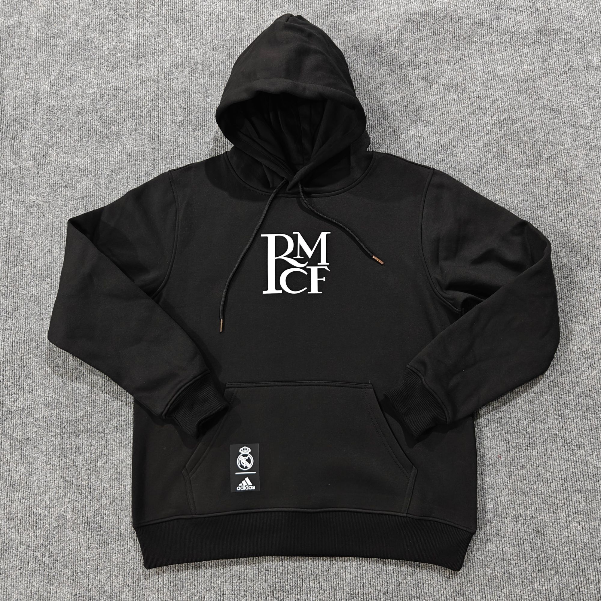 73d0cb5c | UAE SPORT STORE Black hoodie with hood up, white RM graphic on chest, laid on gray carpet; Real Madrid and Adidas tags attached.