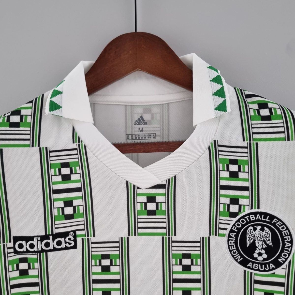 White Adidas polo on a wooden hanger with a bold green–black geometric pattern and a Nigerian Football Federation badge visible on the right.