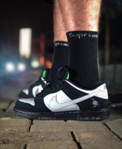 Pair of black-and-silver Nike sneakers worn with tall black socks that say 'Supreme' during night on a brick path.