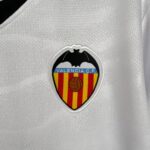 Close-up of the Valencia C.F. crest on a white textured jersey, showing the bat emblem above a blue band and stripes.