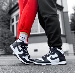 Pair of black-and-white Nike high-top sneakers on a gray pavement, with one person wearing red pants and the other in black joggers.