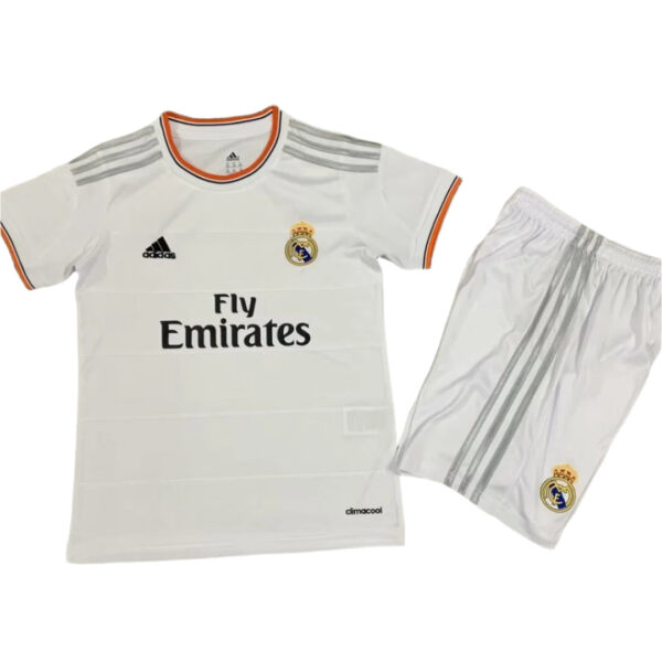 White Real Madrid football jersey with orange trim and crest, featuring the Fly Emirates sponsor, paired with white shorts with gray stripes on the side.