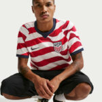Person wearing a USA soccer jersey with red and white diagonal stripes, sitting against a light background, hands together in front.