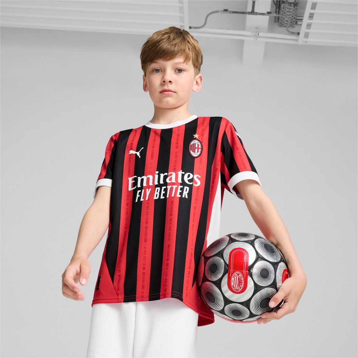 Boy wearing a red-and-black AC Milan jersey holds a patterned soccer ball against a gray backdrop, looking at the camera.