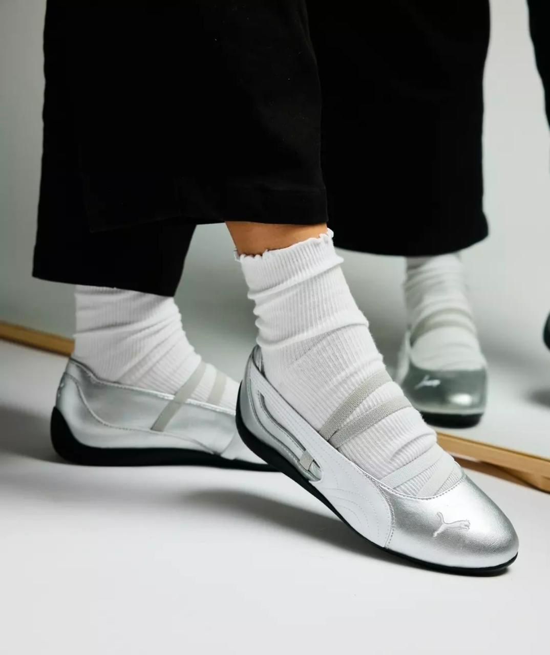 77770057-0c6c-416b-a005-b22fb6588f82 | UAE SPORT STORE Person wearing white socks and silver Puma sneakers stepping forward, with a mirrored background and black pants in view.