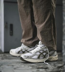 White and gray athletic sneakers with laces on a concrete floor, brown pants visible at the ankles.