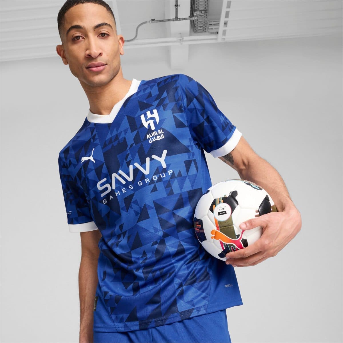 Male athlete in a blue geometric-pattern soccer jersey holding a soccer ball, indoors.