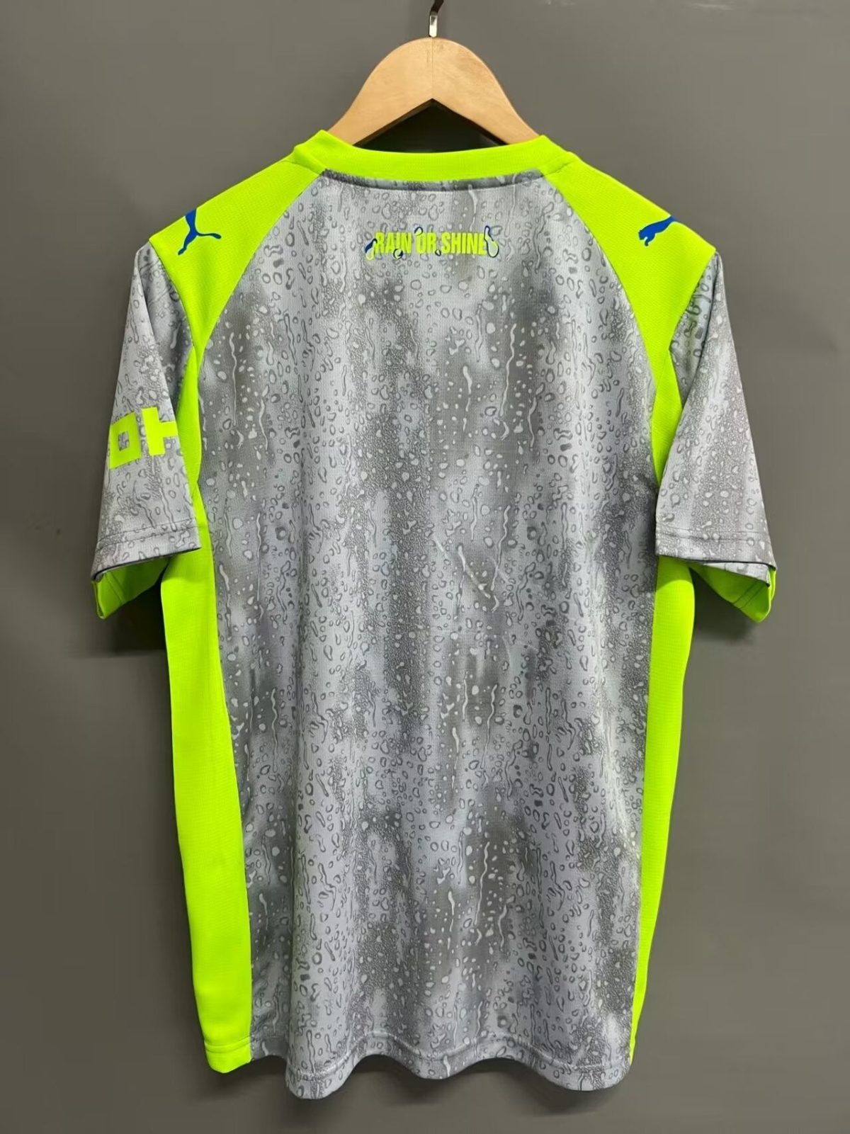 Back of a gray rain‑pattern jersey with neon yellow panels, hanging on a wooden hanger; 'RAIN OR SHINE' text on upper back.